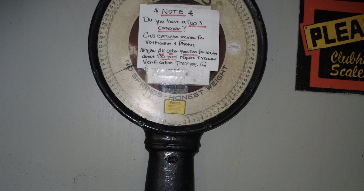 A vintage mechanical scale with a circular dial and a note attached, indicating instructions for verification and photo submission.