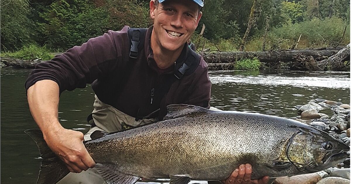 Person holding a large salmon fish while standing in a river, wearing fishing gear and a cap.
